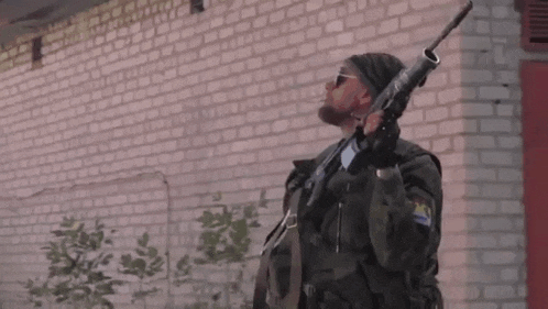 a man with a beard is holding a gun in front of a brick building .