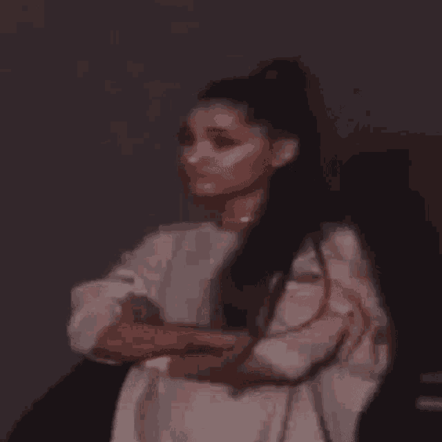 ariana grande is wearing a white jacket and a ponytail while standing with her arms crossed .