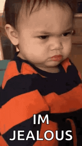 a baby is making an angry face with his arms crossed and the words `` im jealous '' .