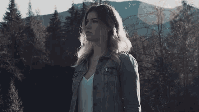 a woman in a denim jacket is standing in the woods
