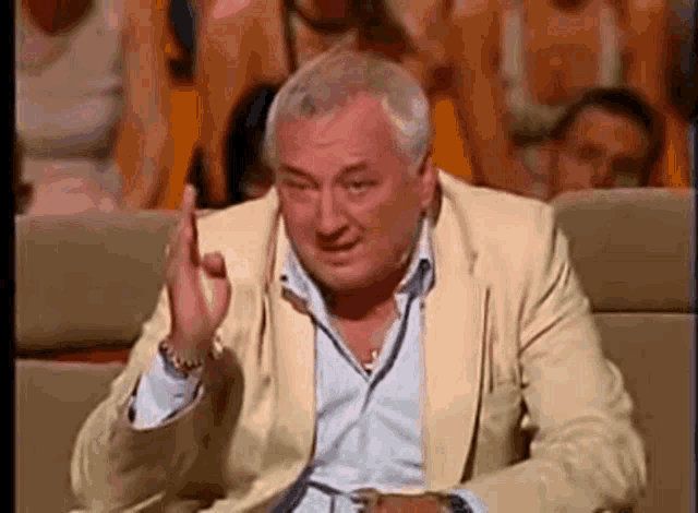 a man in a suit is sitting in front of a crowd and making a gesture with his hand .