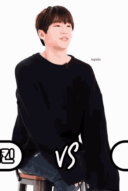 a young man in a black sweater sits on a stool with the words vs on the bottom right