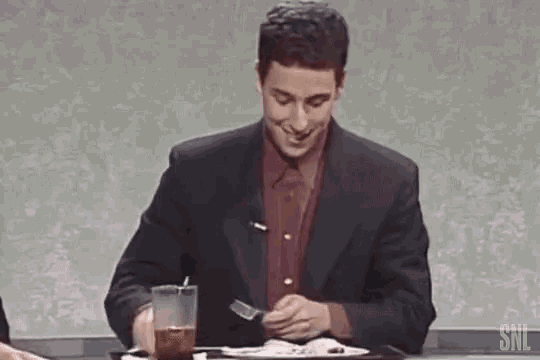 a man in a suit and red shirt is sitting at a table eating food with a fork .