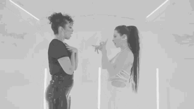 two women are standing next to each other in a room and talking to each other in a black and white photo .