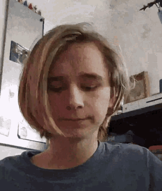 a young boy with long blonde hair is wearing a blue shirt and making a face .