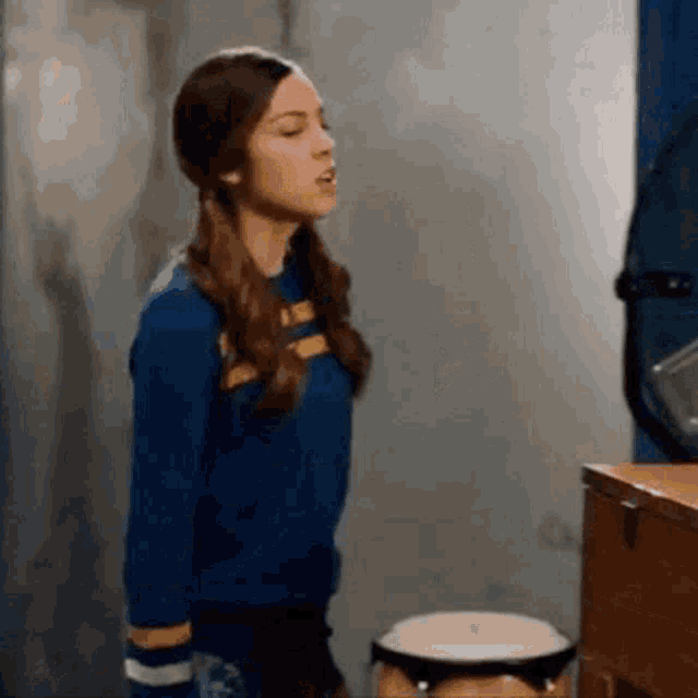 a woman in a blue sweater is standing next to a drum and singing .