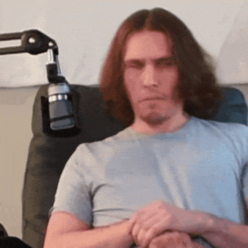 a man with long hair is sitting in a chair with a microphone in the background