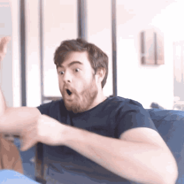 a man with a beard is sitting on a blue couch and making a surprised face