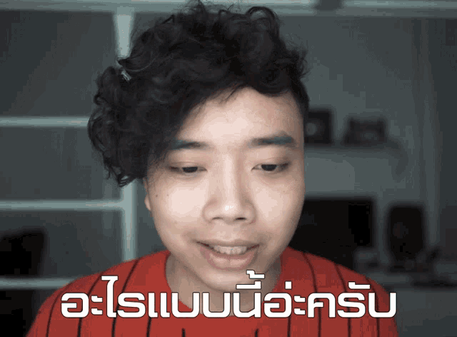 a young man with curly hair is wearing a red and black striped shirt and says " อะไร แบบ นี้ "