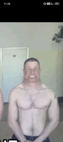 a man without a shirt is standing in front of a door with the time of 11:26 on his phone