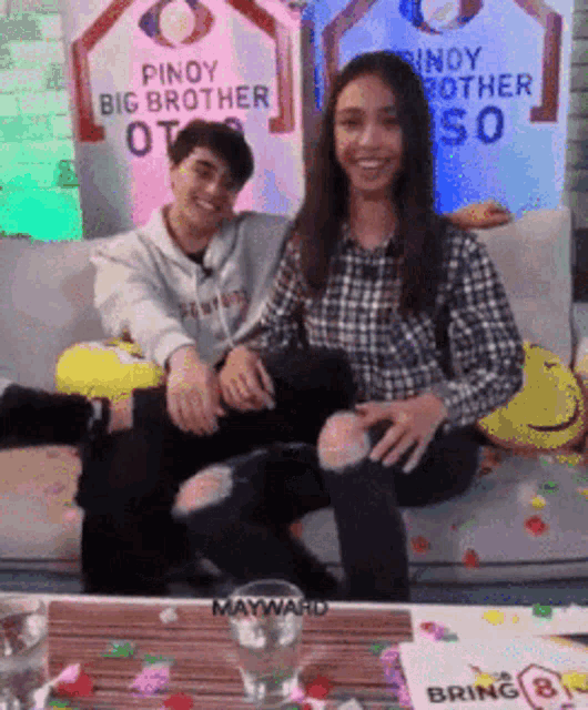 a man and a woman are sitting on a couch with a pinoy big brother sign behind them