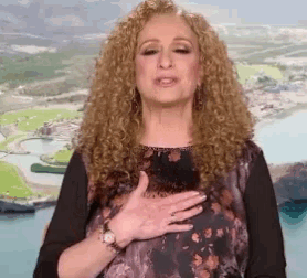a woman with curly hair is holding her hands to her chest in front of an aerial view of a city .