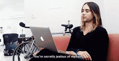 a man with long hair is sitting on a couch using an apple laptop and saying you 're secretly jealous of my hair