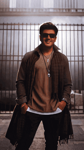 a man wearing sunglasses and a long coat stands in front of a fence
