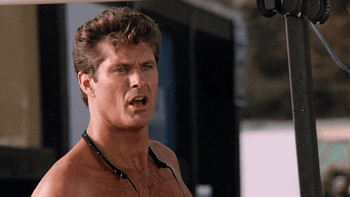 a shirtless man with headphones around his neck looks at the camera with his mouth open