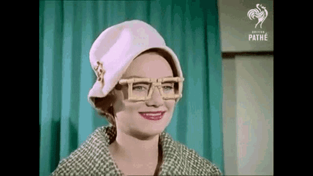 a woman wearing a hat and glasses is smiling in front of a green curtain .