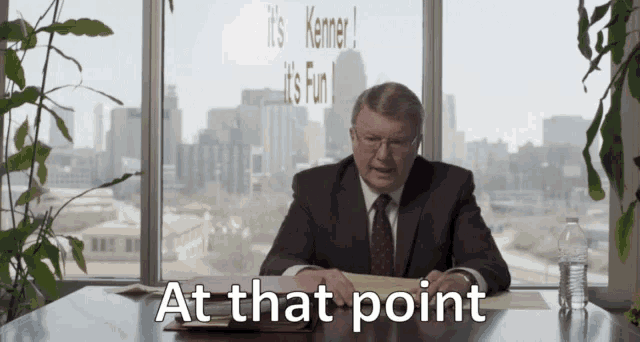 a man in a suit and tie sits at a desk in front of a window with the words at that point above him