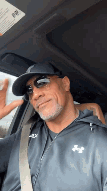 a man wearing sunglasses and a black under armour jacket is sitting in a car