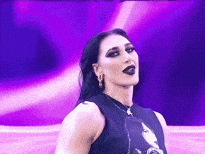 a woman in a black tank top is standing in front of a purple background and looking at the camera .