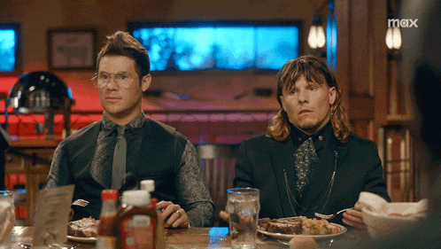 two men are sitting at a table with a max logo on the bottom