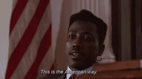 a man in a suit and tie stands in front of an american flag and says " this is the american way "