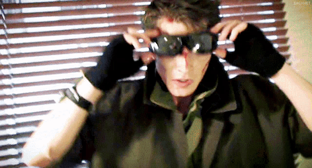a man wearing sunglasses and gloves is standing in front of a window with blinds .