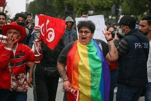 a woman is holding a rainbow flag and a sign while walking down a street with a crowd of people .