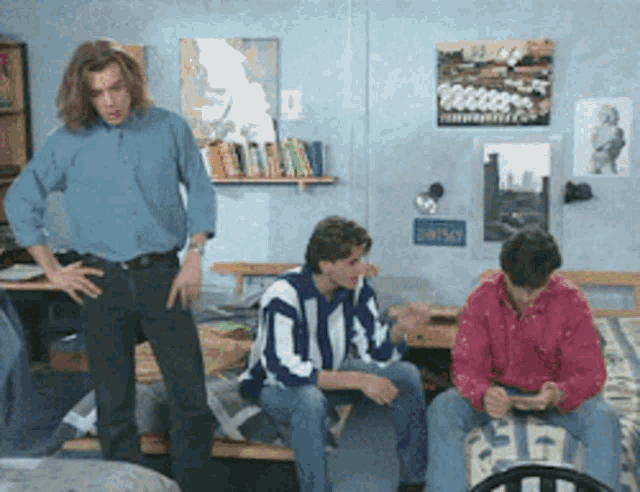 three men in a dorm room with one wearing a blue and white striped shirt with the letter q on the front
