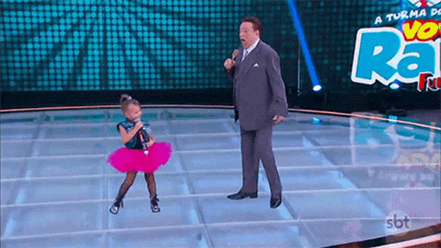 a little girl in a pink tutu is singing into a microphone while a man in a suit stands behind her .