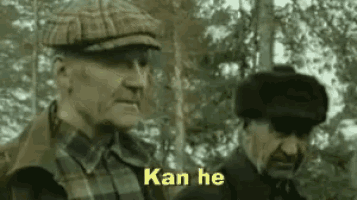 two older men are standing next to each other in the woods and one of them is wearing a hat .