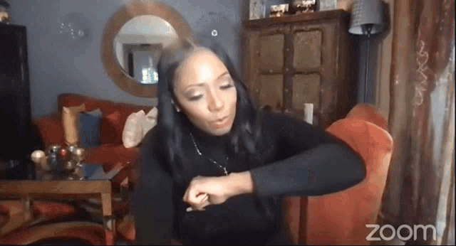 a woman is sitting in a living room on a zoom call looking at her watch