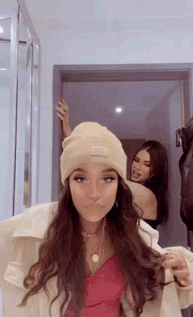 a woman wearing a beanie with the word supreme on it stands next to another woman