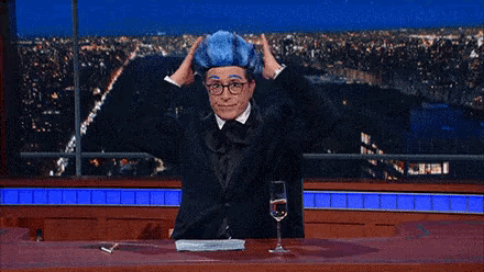 a man wearing a blue wig and glasses is sitting at a table with a glass of wine .