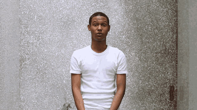 a man in a white t-shirt stands in front of a gray wall