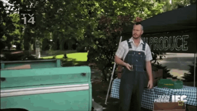 a man wearing overalls stands in front of a produce stand