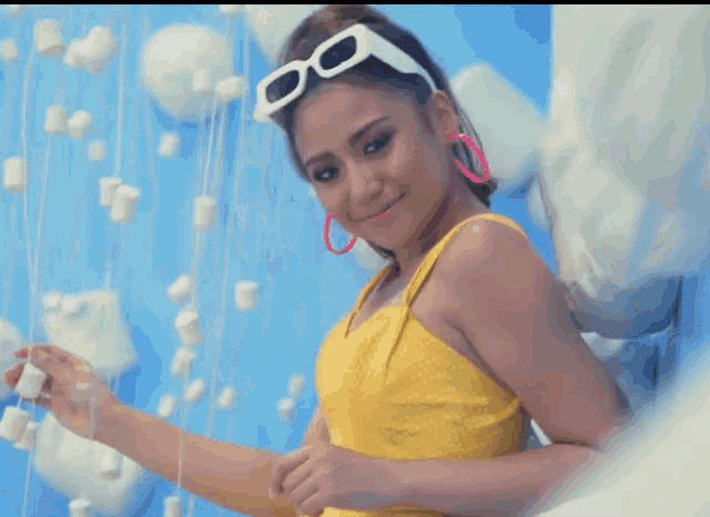 a woman wearing a yellow dress and white sunglasses is standing in front of a wall of marshmallows
