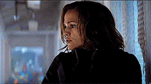 a woman in a black jacket is standing in a dark room looking out a window .