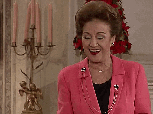 an older woman in a pink jacket and necklace is standing in a living room .