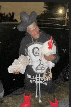 a man wearing a billionaire studios sweatshirt holds a chicken
