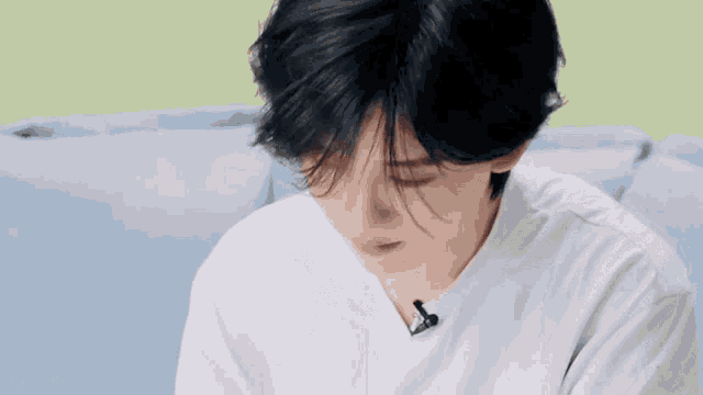 a young man with black hair is sitting on a couch with his hand on his chin .