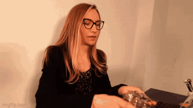 a woman wearing glasses is sitting at a desk holding a bottle of liquid .