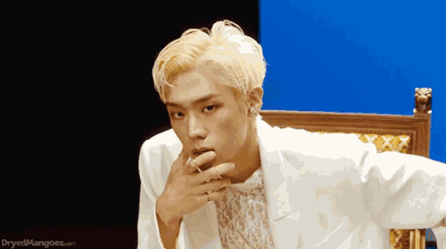 a young man in a white suit is sitting in a chair with his hand on his chin