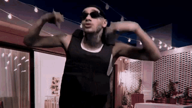 a man wearing sunglasses and a black tank top is dancing in a room with string lights .