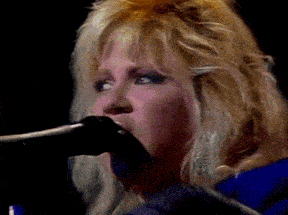 a blonde woman singing into a microphone with a dark background