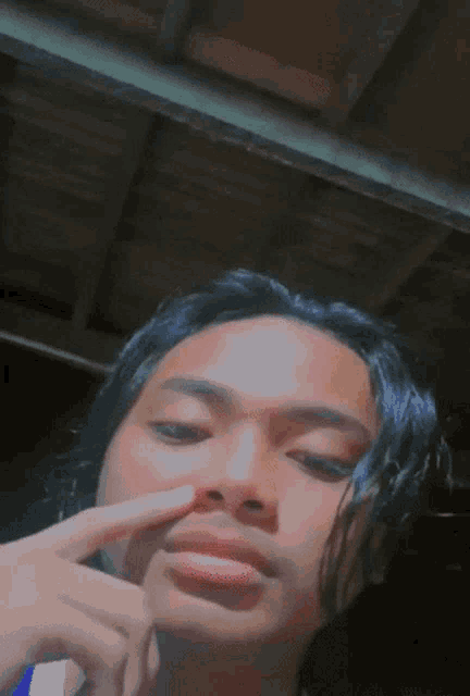 a young man with long black hair is touching his nose with his finger .