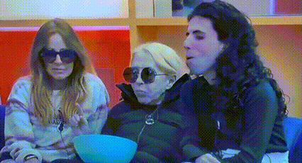 three women are sitting on a couch eating popcorn and one of them is wearing sunglasses .