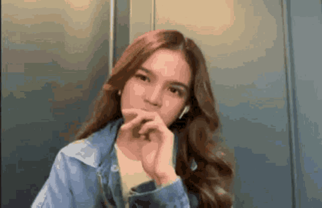 a young woman wearing headphones and a denim jacket is looking at the camera with her hand on her chin .
