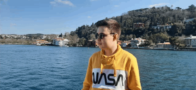 a man wearing sunglasses and a nasa sweatshirt stands in front of a body of water