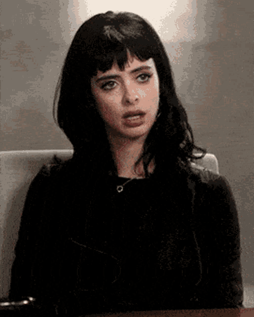 a woman with black hair and bangs is sitting at a table making a funny face .