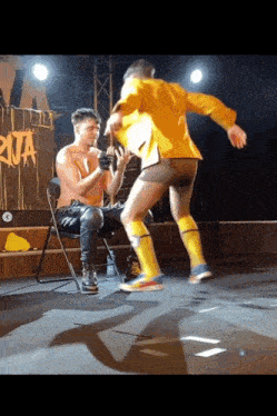 a man in a yellow jacket is standing next to a man sitting in a chair with the word riva on the wall behind him